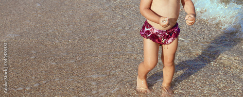 little girl in purple swimming trunks on the beach carries sea pebbles and seashells in her hands. Rest and travel. Vacation concept with little children. People and advertising concept. Childhood. 