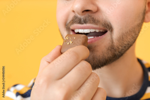 Handsome young man eating tasty chocolate on color background, closeup
