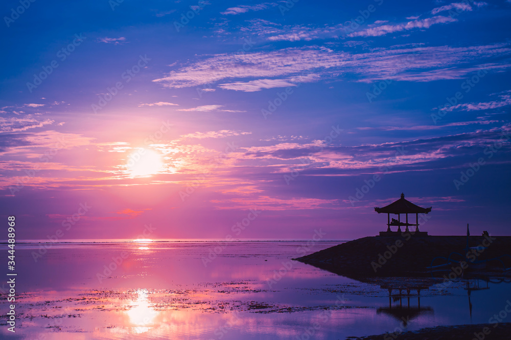 Pink dawn the sun and the gazebo at Sanur beach, Bali, Indonesia. A calm surface of sea water that reflects the sun and cloudy sky. Pink dawn the sun and the gazebo at Sanur beach, Bali, Indonesia. A calm surface of sea water that reflects the sun and cloudy sky.