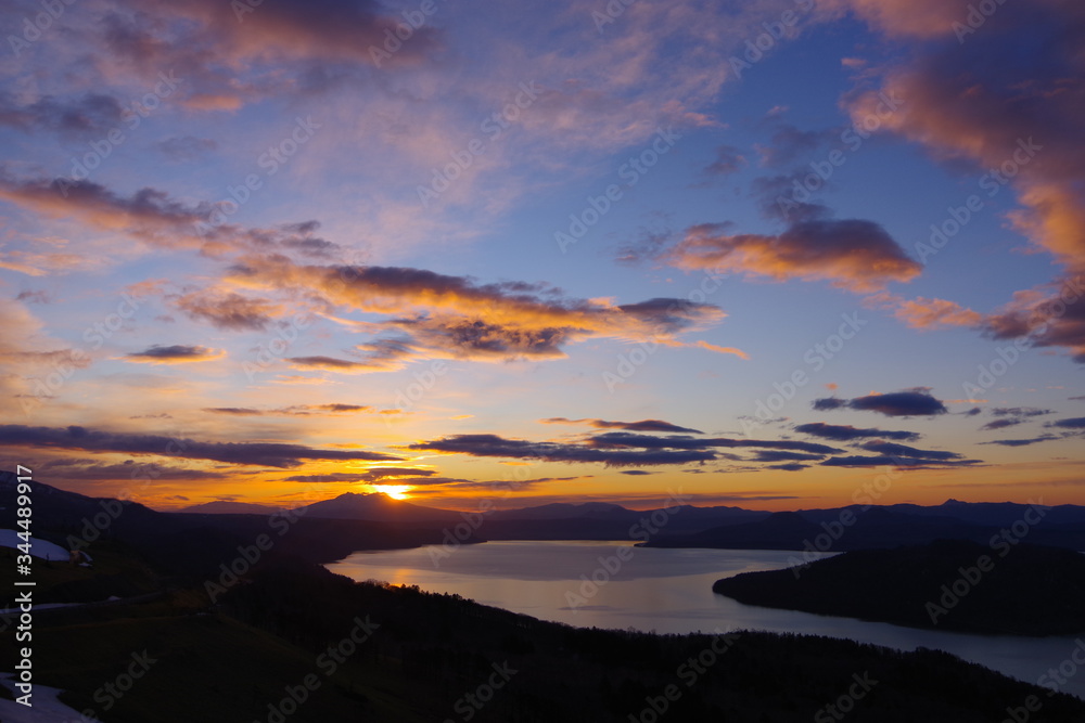 峰から望む夜明けの空のグラデーション。朝の太陽に照らされた雲。眼下の湖。北海道、日本。