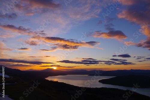 峰から望む夜明けの空のグラデーション。朝の太陽に照らされた雲。眼下の湖。北海道、日本。