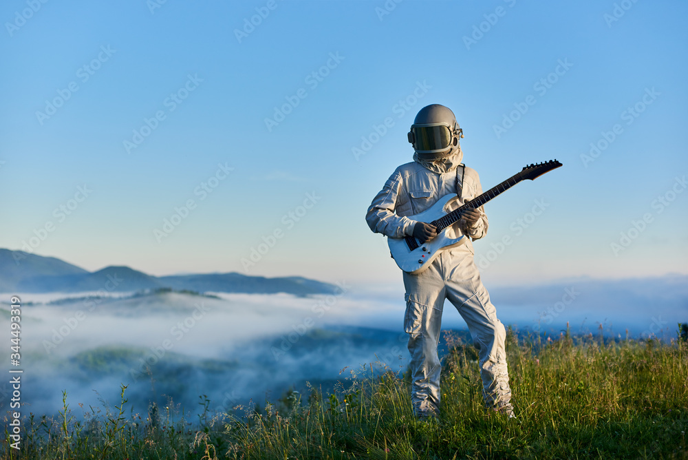 Astronaut wearing white space suit and helmet playing white guitar, standing on sunny green mountain glade in summer under blue sky, morning fog rising up from the valley behind him. Astronaut wearing white space suit and helmet playing white guitar, standing on sunny green mountain glade in summer under blue sky, morning fog rising up from the valley behind him.