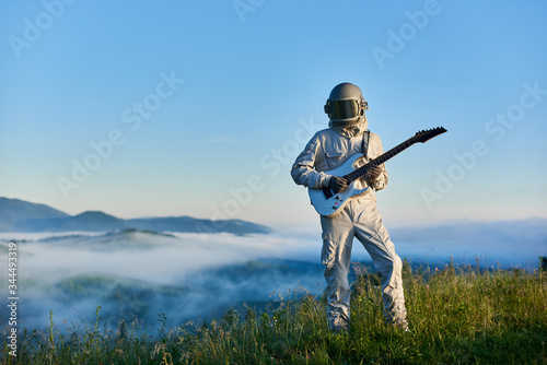 Astronaut wearing white space suit and helmet playing white guitar, standing on sunny green mountain glade in summer under blue sky, morning fog rising up from the valley behind him.