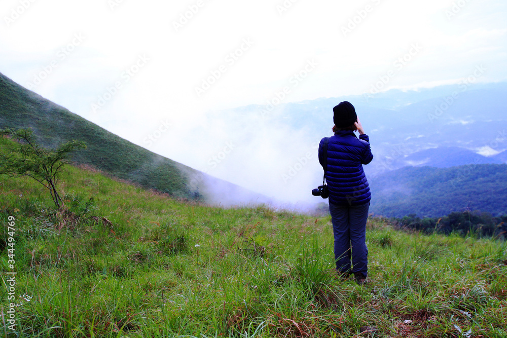 Naklejka premium Asian hiking or tourist woman in dark blue sweater with camera standing on green grass field with mountain view and mist background and copy space. Beautiful in nature and Landscape
