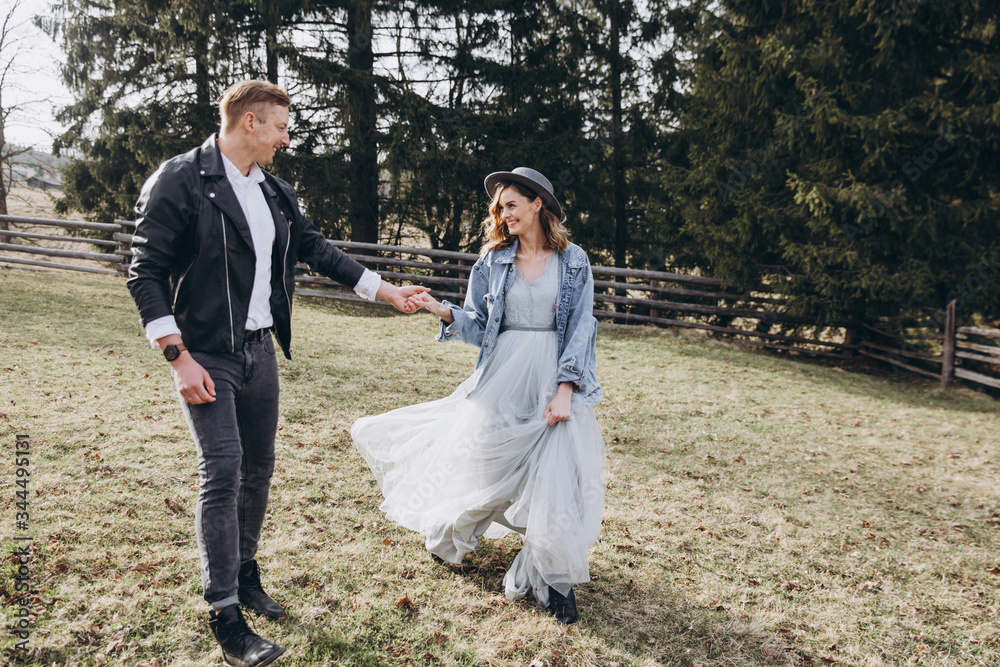 Country wedding. A guy in a leather jacket and a young girl in a gray-blue dress and hat walk along a clearing in the mountains against the background of a forest and a wooden fence at sunset Country wedding. A guy in a leather jacket and a young girl in a gray-blue dress and hat walk along a clearing in the mountains against the background of a forest and a wooden fence at sunset