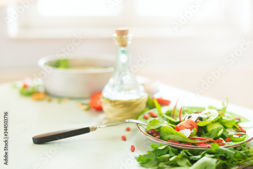 Healthy cuisine. Full bowl of fresh salad served on light kitchen table with olives oil and fork at sunny window background. Diet or vegetarian food concept. Home food eating