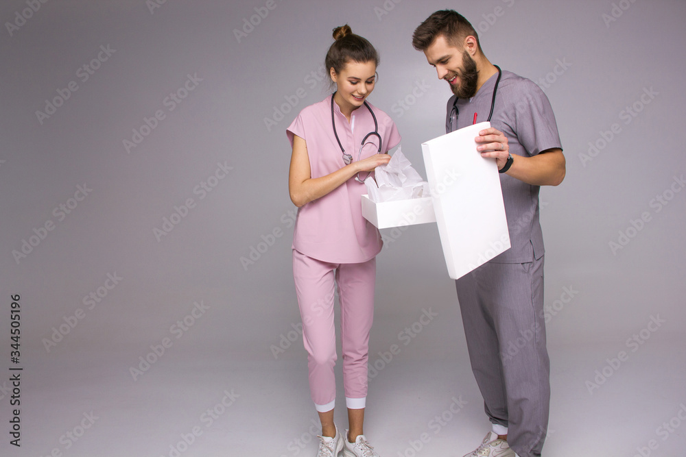 doctors relationship. happy male and female nurses in medical costumes with stethoscopes on neck are unpacking white box with emotions on the gray background, lifestyle medical concept, free space doctors relationship. happy male and female nurses in medical costumes with stethoscopes on neck are unpacking white box with emotions on the gray background, lifestyle medical concept, free space