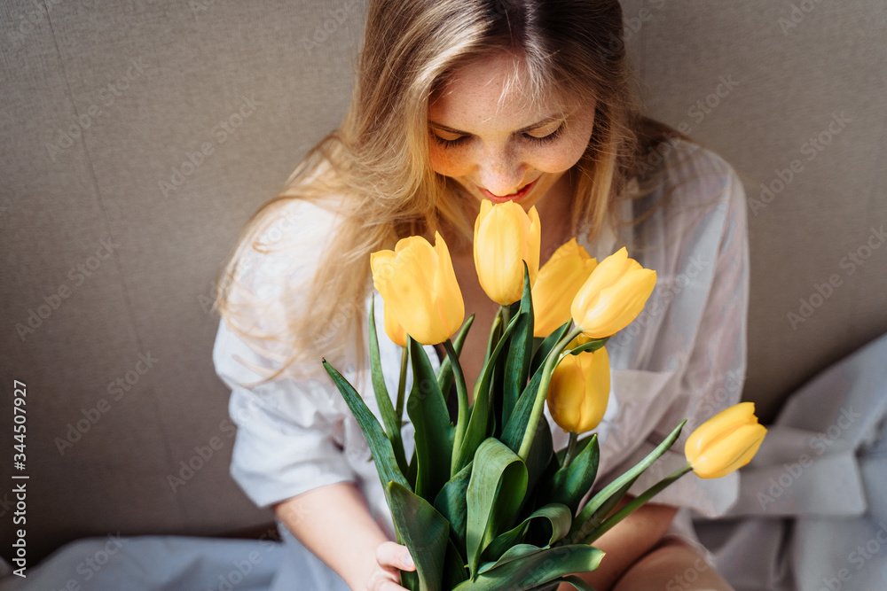 Beautiful young blond woman with tulip bouquet. Close-up spring portrait. Happy and romantic woman at home interior with sun rays and a bouquet of yellow flowers. Girl in underwear and white shirt