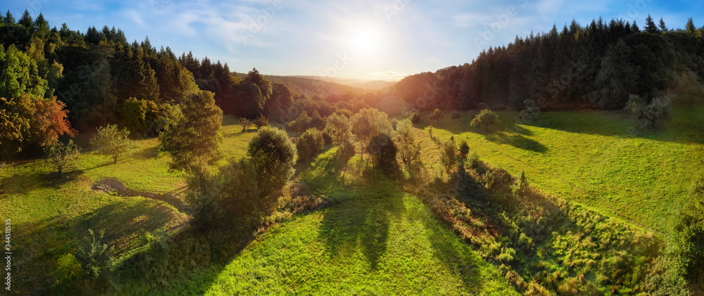 Aerial landscape panorama after sunrise: gorgeous scenery with the sun in the blue sky, trees on green meadows casting long shadows, surrounded by forests on hills