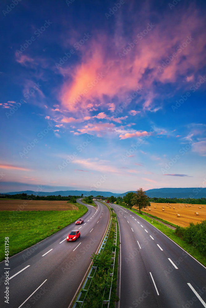 Wide, almost empty road under the colorful sky after sunset, with beautiful red clouds and two red cars with slight motion blur, a dynamic transportation and landscape shot