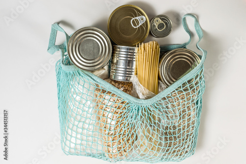 various long-term storage products in tin cans, spaghetti paste, sugar, dry peas and pearl barley in bundles in a blue mesh bag on a white background