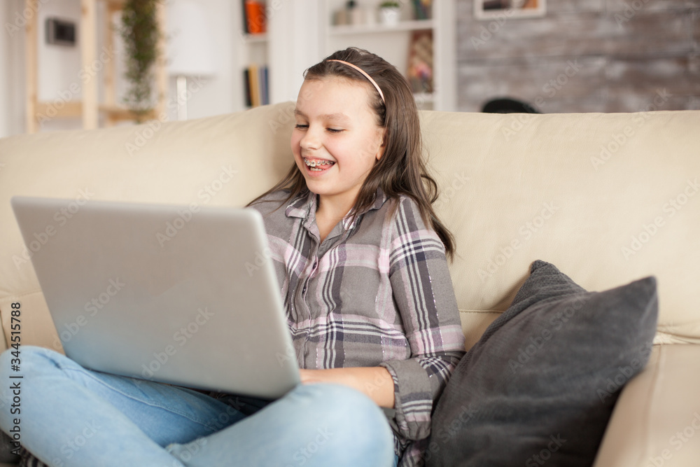Fototapeta premium Smiling little girl with braces using her laptop in living room. Cheerful child.