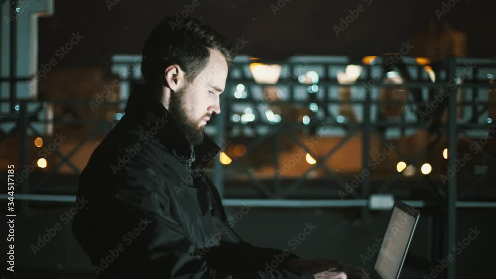 guy with a beard hipster at night sits on the roof with a laptop and works remotely in the night lights guy with a beard hipster at night sits on the roof with a laptop and works remotely in the night lights
