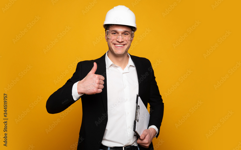 Good work! Positive foreman in a suit and helmet is looking in the camera and showing thumbs up, while holding his notes and smiling. Good work! Positive foreman in a suit and helmet is looking in the camera and showing thumbs up, while holding his notes and smiling.