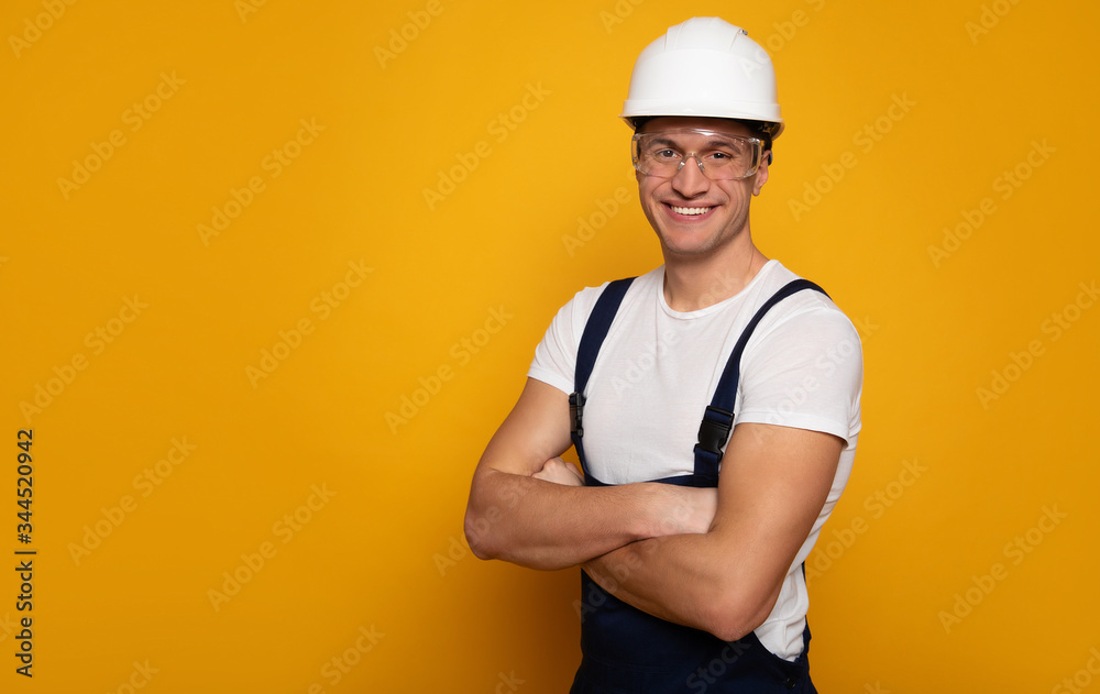 Friendly builder. Close-up photo of a young man in a working clothes, who is wearing a helmet and transparent glasses, while standing with his arms folded.