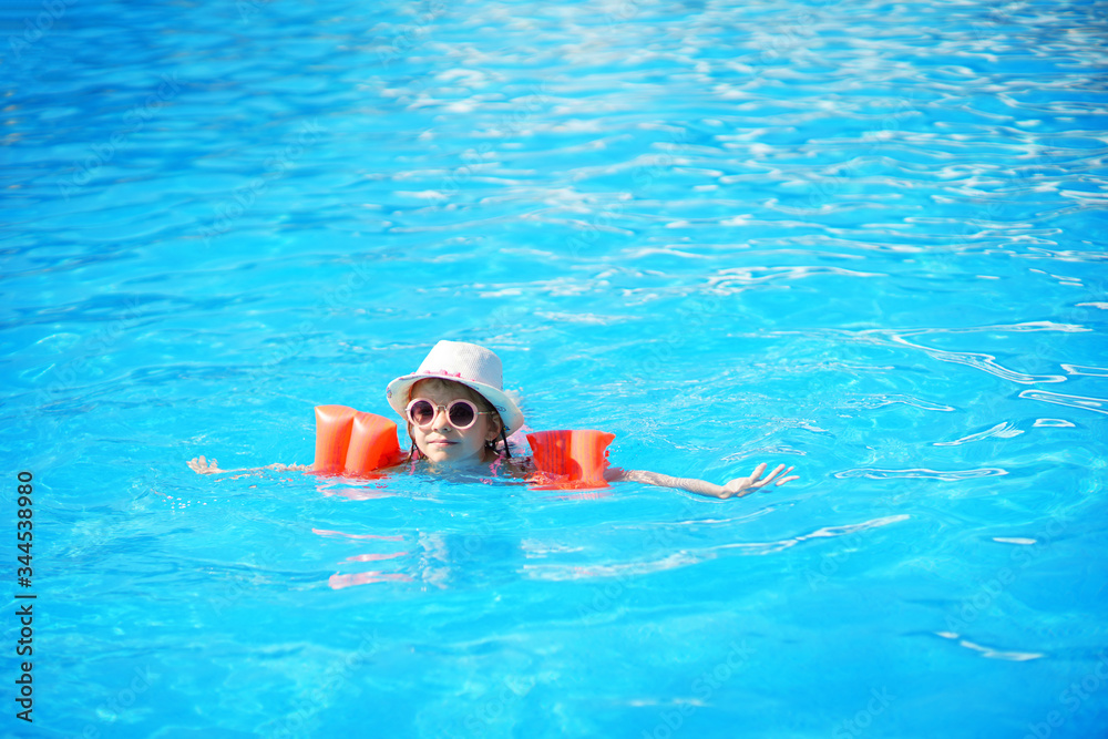 A charming child of European appearance, a girl 7-8 years old, swimming in the pool. Summer, a clear and sunny day. A girl with her family on vacation at sea. The girl has inflatable arm ruffles.