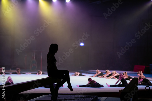 A young female gymnast is sitting on a gymnastics beam ready to perform her routine while waiting for the younger gymnast to finish on the floor. The spotlights illuminate the floor but not the girl.