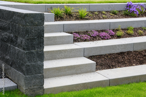 Moderne terrassenförmige Gestaltung eines Garten  Hanggrundstücks: Treppe und Stufen aus Betonsteinen mit Gräsern und lila Blumen geschützt mit Mulch und begrenzt von einer grauen Naturstein Mauer  photo