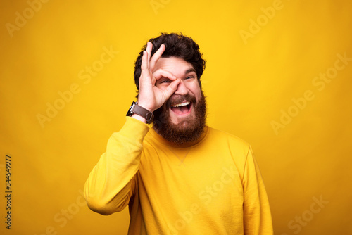 Playful bearded man is looking at the camera through an ok gesture on his eye on yellow background.