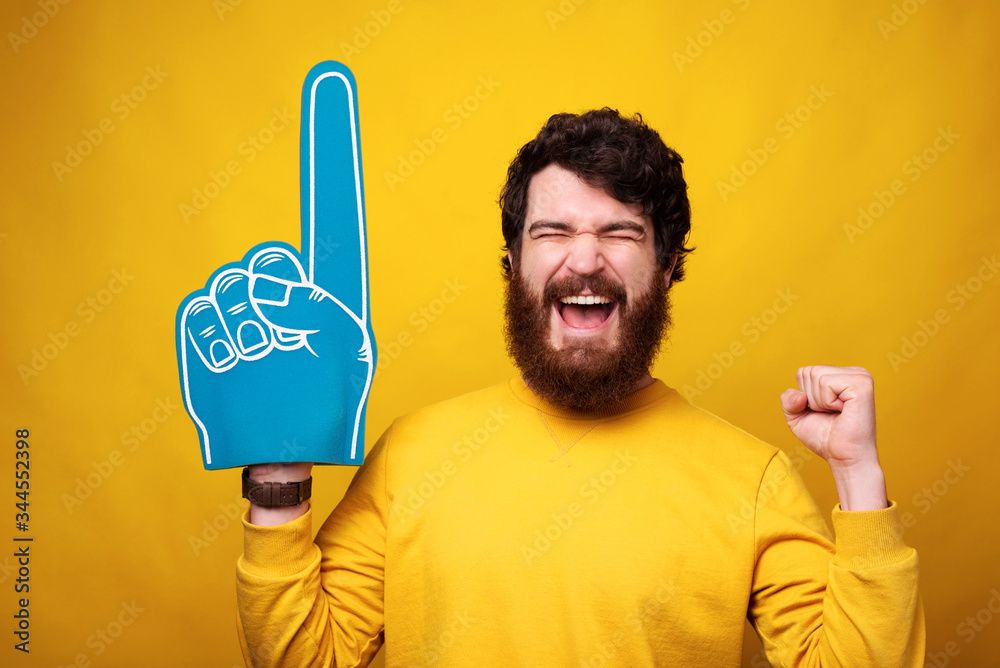Excited young man is wearing a foam fan glove while making winner gesture  on yellow background. Stock-Foto | Adobe Stock