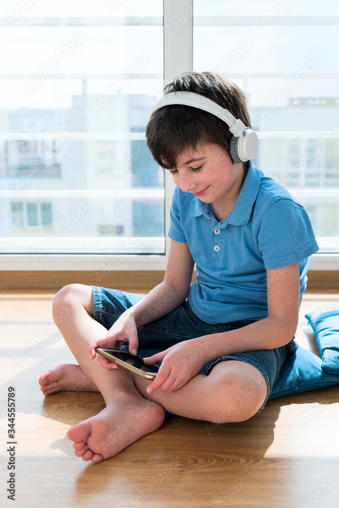 Nine years old child wearing headphones using smartphone sitting on pillow. Soft focus. Quarantine and online school concept