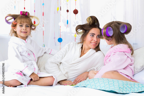 Young woman lying on the bed with the two daughters and curlers in hair at home. Stay home and family concept.
