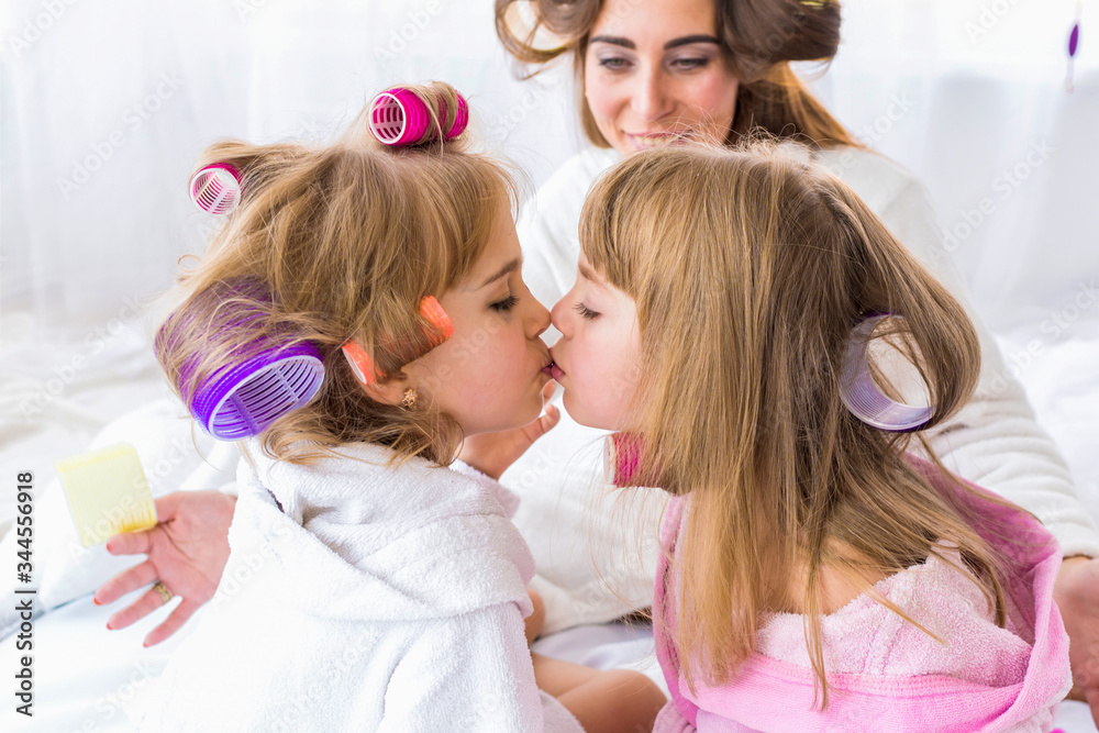 Two daughters are kissing with curlers in hair while mom looks at them on the bed in the bedroom. Stay home and family concept.