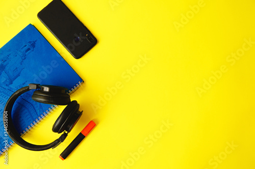 A large blue notebook, headphones, smartphone and marker on a yellow background. Flat layout with space for text