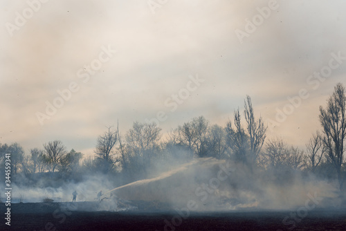 Raging forest spring fires. Burning dry grass, reed along lake. Grass is burning in meadow. Ecological catastrophy. Fire and smoke destroy all life. Firefighters extinguish Big fire. Lot of smoke photo