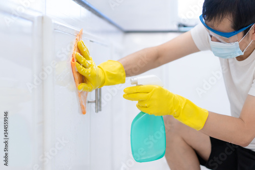 Asian young man wear the protective gloves, glasses and mask is cleaning the surface cabinet in the kitchen with spray cleaner and wet clothes, concept of hygiene concern in covid 19 virus outbreak.