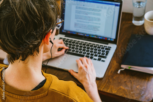 Woman working on desk in home office in old vintage room. Young Caucasian short hair woman with earphones working online from home on computer laptop from back, head and shoulders, real, candid photo 