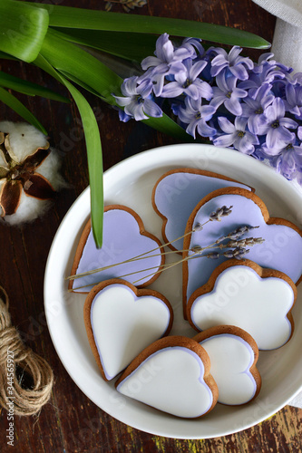 Rustic flat lay. Heart-shaped white and lavender gingerbread lies in white plate on dark wood table with cotton flower and purple hyacinth flower . Saint Valentine's day.