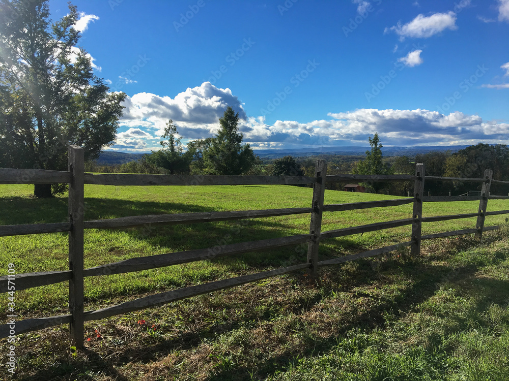 Scenic view with wooden fence in foreground and mountains and trees in the background brilliant blue sky and puffy clouds with strong shadows