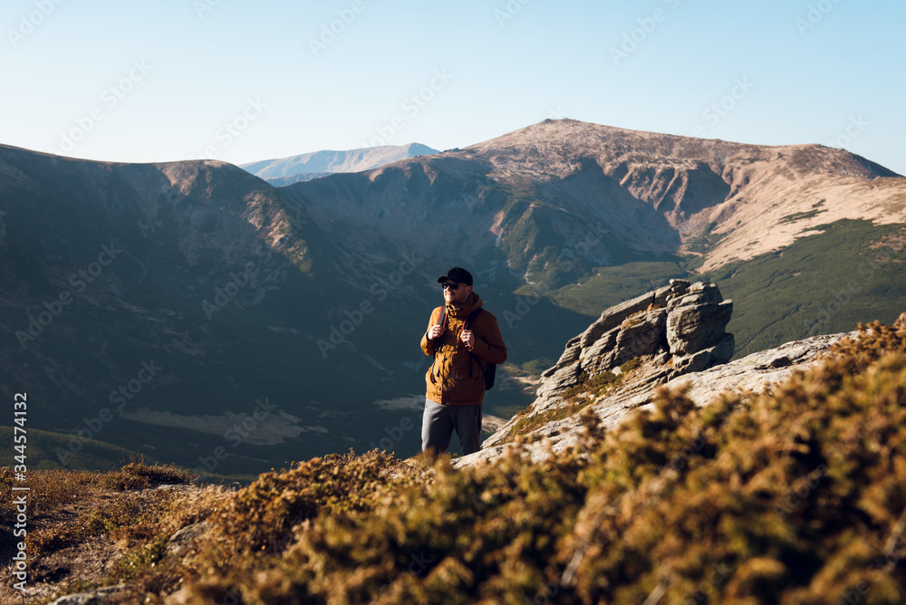 Young man in cap and sunglasses with a backpack walks the trail at the top of the mountain. Mountain hike in the sunny Carpathians. Hiking concept Young man in cap and sunglasses with a backpack walks the trail at the top of the mountain. Mountain hike in the sunny Carpathians. Hiking concept