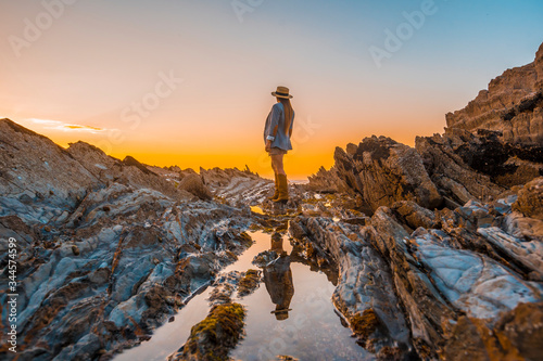 Lifestyle, a young businesswoman on the rocks by the sea with jacket and hat reflected in the water