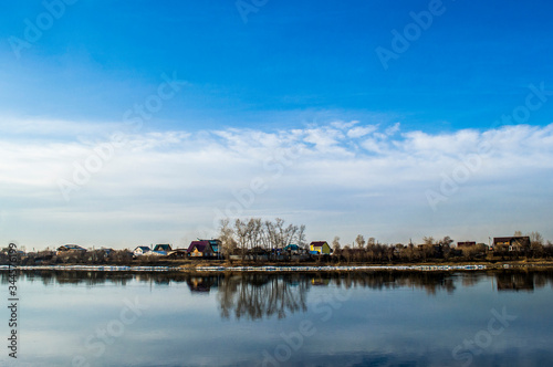 Horizon dividing a blue sky with clouds and a river with reflection of the sky and houses © GhosterzArts