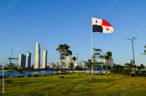 La bandera de Panamá izada en el mástil, ondeando en el viento, en un espacio abierto cubierto de hierba, detrás del Océano Pacífico y la vista de los altos edificios de la ciudad.