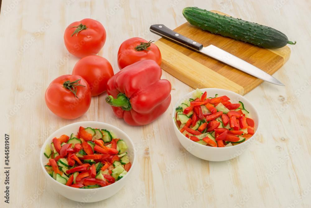 White bowls with fresh salad of green cucumbers, red tomatoes and bell peppers on light wooden surface. Vegetables. Lowcalorie vitamin dietary food. Concept of healthy lifestyle White bowls with fresh salad of green cucumbers, red tomatoes and bell peppers on light wooden surface. Vegetables. Lowcalorie vitamin dietary food. Concept of healthy lifestyle