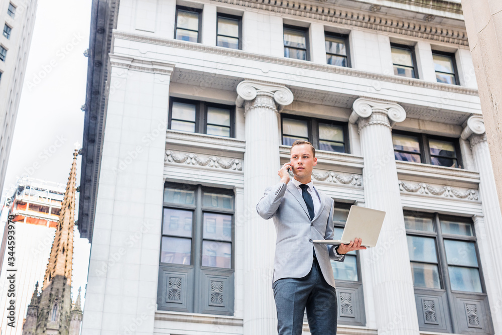Young Businessman traveling, working in New York, wearing gray blazer, white shirt, black tie, standing on street outside vintage office building, working on laptop computer, talking on cell phone.. Young Businessman traveling, working in New York, wearing gray blazer, white shirt, black tie, standing on street outside vintage office building, working on laptop computer, talking on cell phone..
