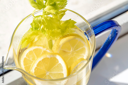 Lush green-yellow lemon slices and celery leaves in a blue handle glass carafe next to window at sunlight with high PH level detox water to boost the immune system to fight corona virus. 