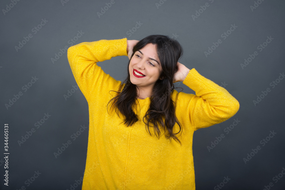 Confidence and coquettish concept. Portrait of charming young girl, smiling broadly with self-assured expression while holding hands over her head. Standing against gray background. Confidence and coquettish concept. Portrait of charming young girl, smiling broadly with self-assured expression while holding hands over her head. Standing against gray background.