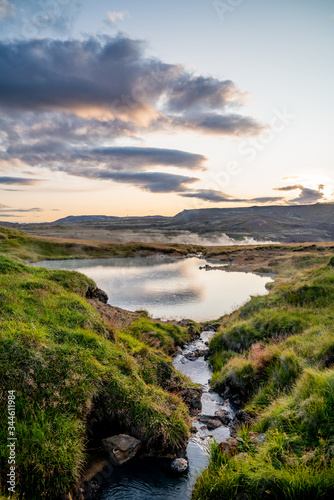 A small creek leading up to a little lake in a green environment. The stream passes over a few rocks. In the distance there is smoke from a geothermal spring and the sun setting below the mountains.