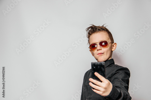 Photos a fashionable guy in sunglasses and a black shirt with a cool hairstyle looks at the phone and photographs himself on a white background