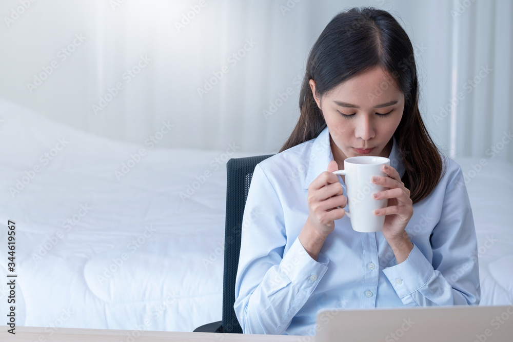 Front view of pretty business girl with blue shirt drink coffee during work at home in bedroom with morning light.