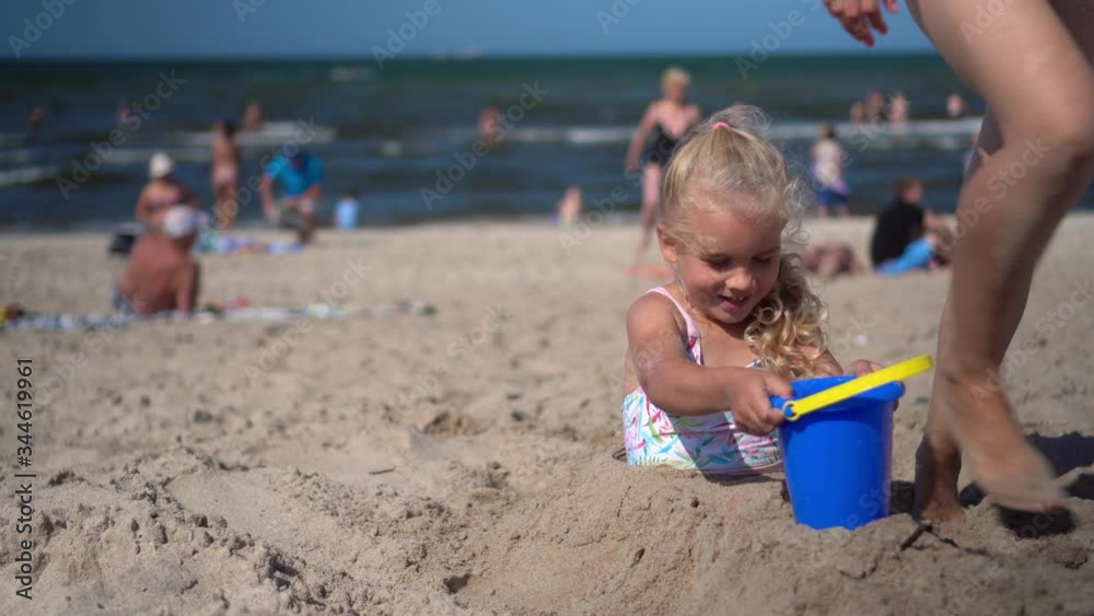 Mother bring daughter buried in sand bucket with water. Blurred people on beach Mother bring daughter buried in sand bucket with water. Blurred people on beach