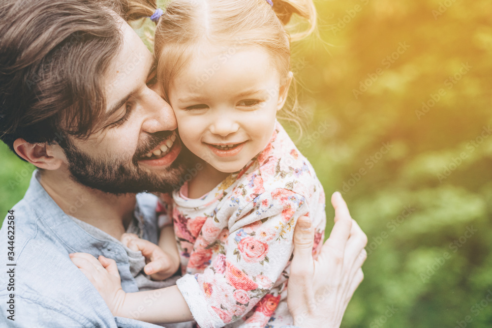 Fototapeta premium stylish bearded father with his little daughter in his arms against the backdrop of green trees on a walk in the park hugging and kissing a baby