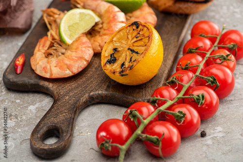 close up view of fried shrimps with lemon and lime near cherry tomatoes on wooden board on grey concrete background