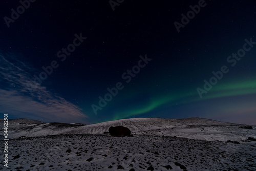Northern lights in Iceland on dark blue sky with a hint of clouds over a snow covered barren mountain landscape with a large rock in the foreground.