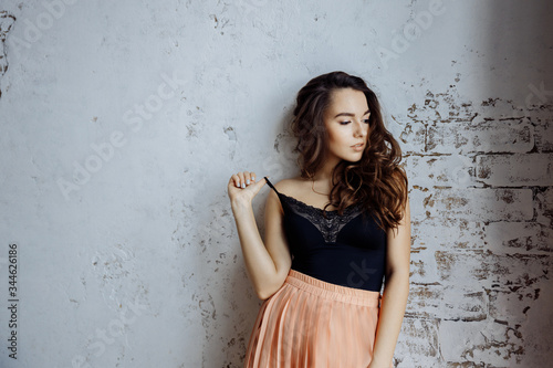 A beautiful girl stands against the wall and looks away. The girl's hand adjusts the strap on the black top. She's wearing a high-waisted pleated skirt.