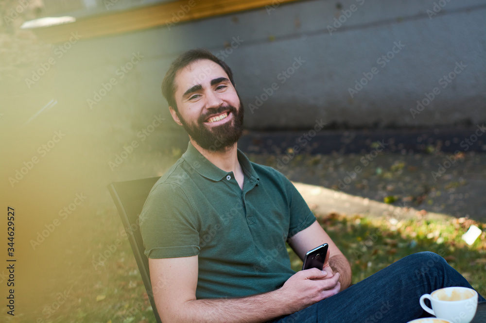 Obraz premium Portrait of young guy with dark beard, sitting in coffee shop on summer terass and drinking coffee in green shirt.